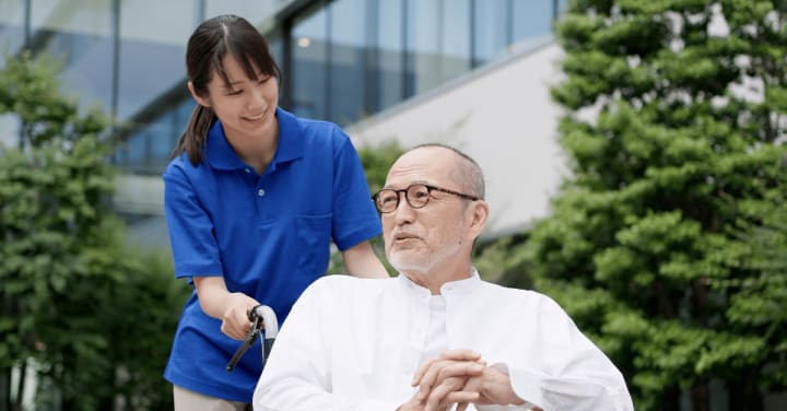 A Smiling Caregiver In A Blue Polo Shirt Walks Outdoors With An Older Man Sitting In A Wheelchair.