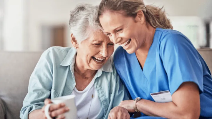 A caregiver in blue scrubs and an older woman share a joyful laugh together indoors.