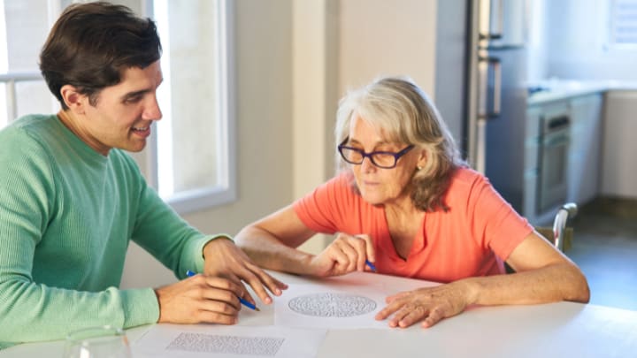 A Young Man In A Green Sweater Sits At A Table And Helps An Older Woman Complete A Paper Maze Puzzle.
