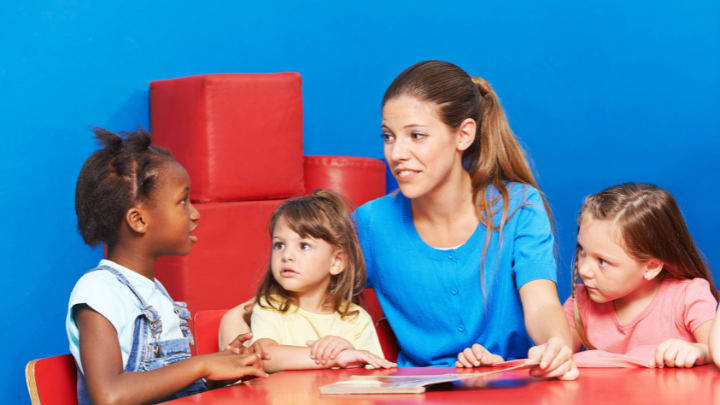 Effective Communication Strategies: Talking To Children About Dementia In Dallas, Tx 1 A Teacher Or Caregiver Sitting At A Table With Three Young Children, Listening Attentively During A Group Activity Or Discussion.