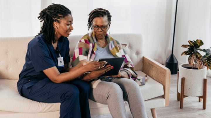 A Caregiver In Dark Blue Scrubs Sits On A Couch And Helps An Older Woman Use A Digital Tablet.