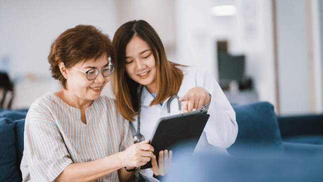 A Healthcare Worker In A White Lab Coat Shows An Older Woman Information On A Digital Tablet.