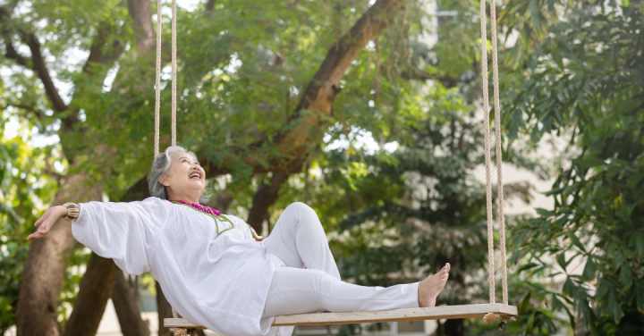 An Older Woman In White Clothing Smiles Joyfully While Leaning Back On A Large Wooden Rope Swing Surrounded By Green Trees.