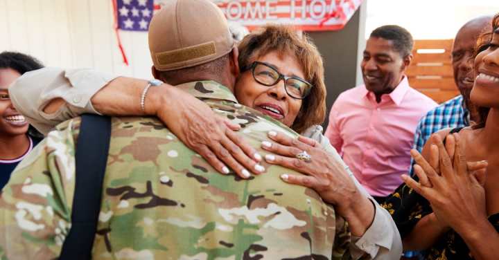 Memory Care: Veteran Dementia Support In Park Cities 1 A Smiling Older Black Woman Hugging A Military Veteran In Uniform As Family Members Look On Joyfully.