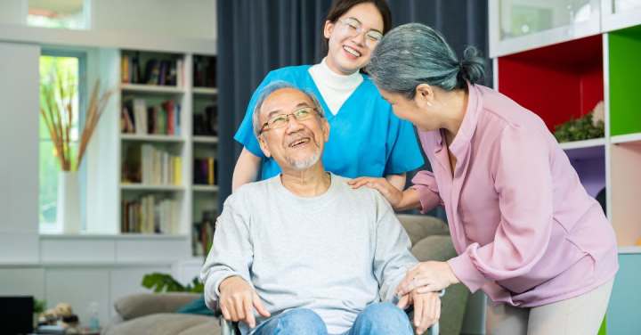 A Smiling Caregiver In Blue Scrubs And An Older Woman Support An Elderly Man Seated In A Chair As They All Share A Warm, Joyful Moment.