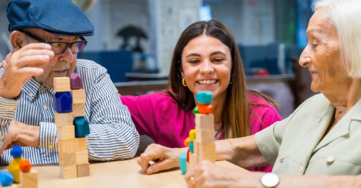A Smiling Caregiver Assists An Older Man And Woman Playing A Tabletop Block Stacking Game.