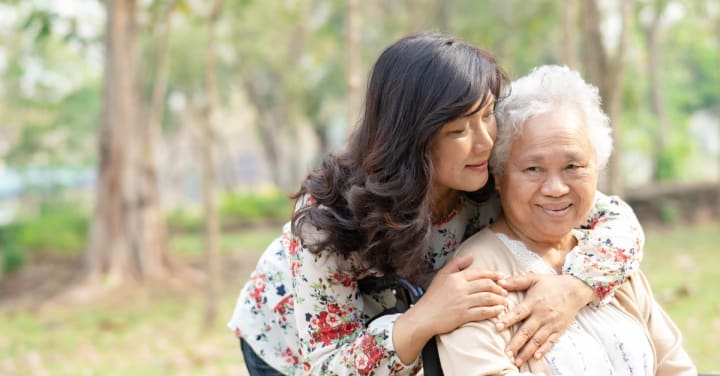 A Younger Woman Gently Embraces An Older Woman From Behind As They Pose Together Outdoors.