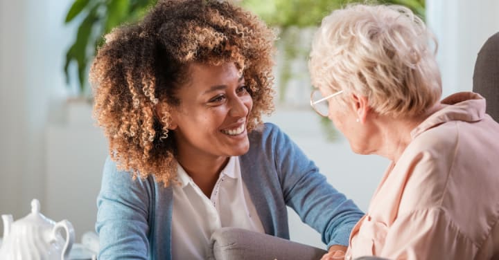 The Essential Guide: What To Look For In Memory Care? 1 A Smiling Younger Woman Looks Warmly At An Older Woman During A Conversation Indoors.