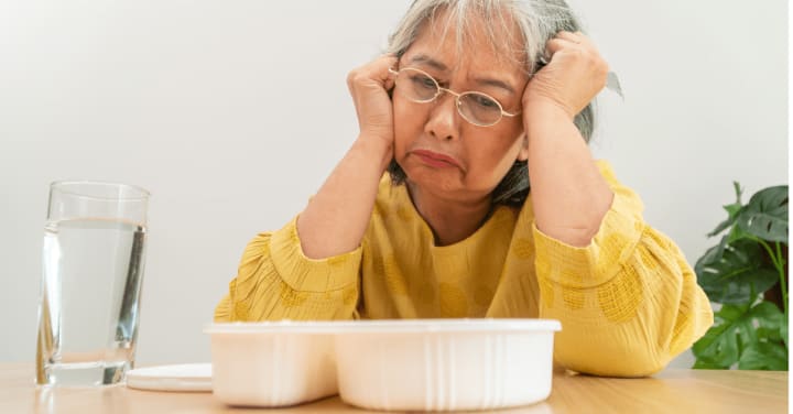 An Older Woman Looks Down With A Sad, Reluctant Expression At A Tray Of Food On The Table In Front Of Her.
