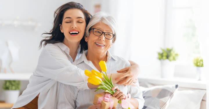 A Younger Woman Joyfully Hugs An Older Woman From Behind As They Smile Together, With The Older Woman Holding A Small Gift And Yellow Tulips.